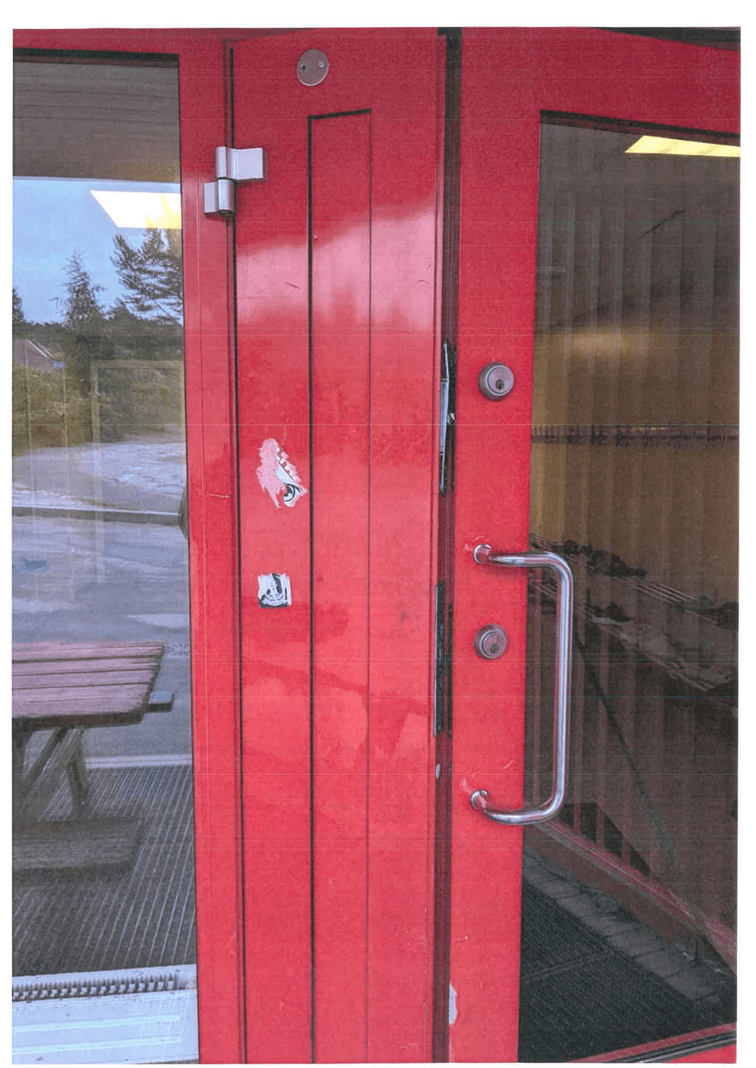 Rød inngangsdør i to fløyer på skolebygg, fotografert utenfra, med stålhåndtak og to låssylindre.