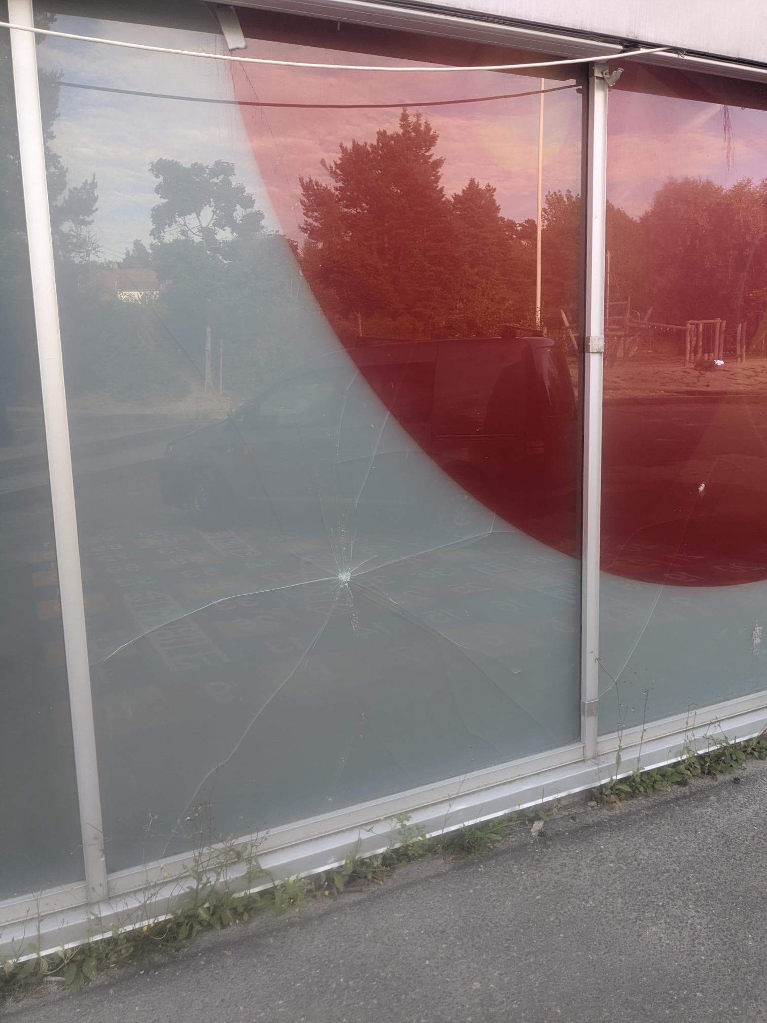 Nærbilde av et sprukket glassfelt i fasaden ved gymsalen på Rød skole, med skolegård og lekeapparater speilet i glasset.