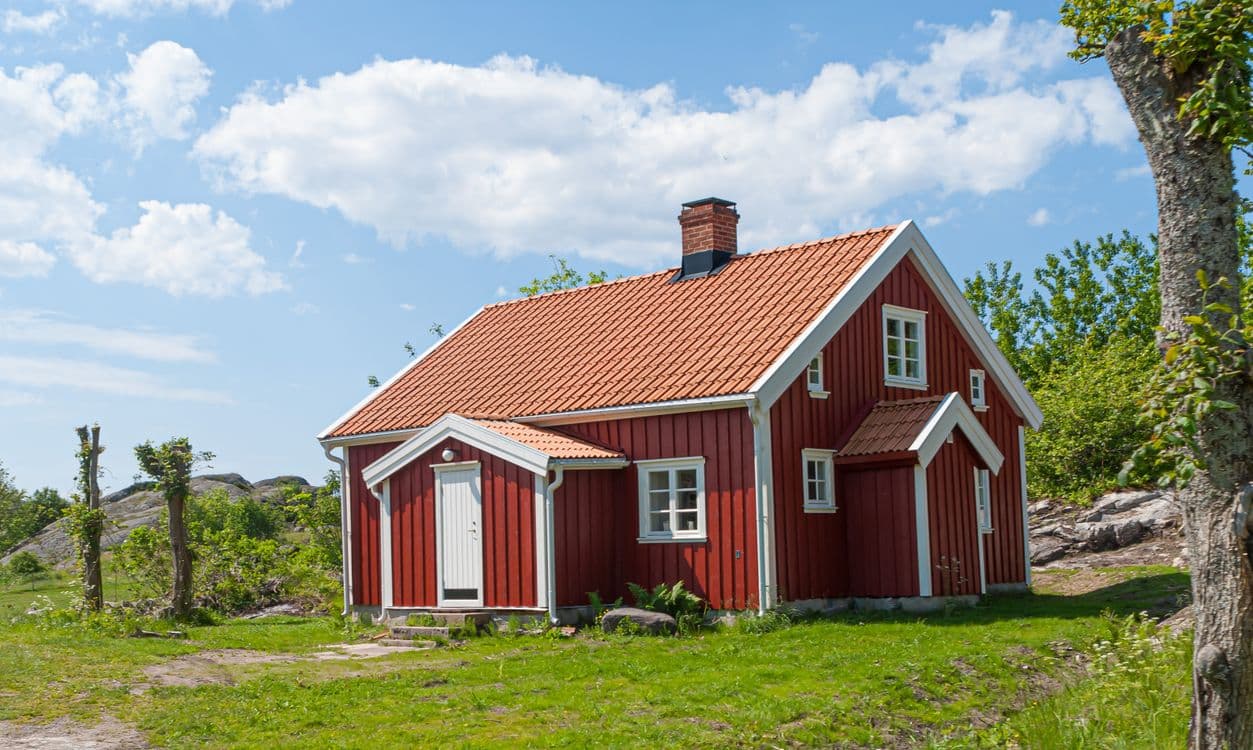Rødt tradisjonelt trehus med hvite vinduskarmer og tegltak på en gresskledd knaus med fjell og blå himmel.